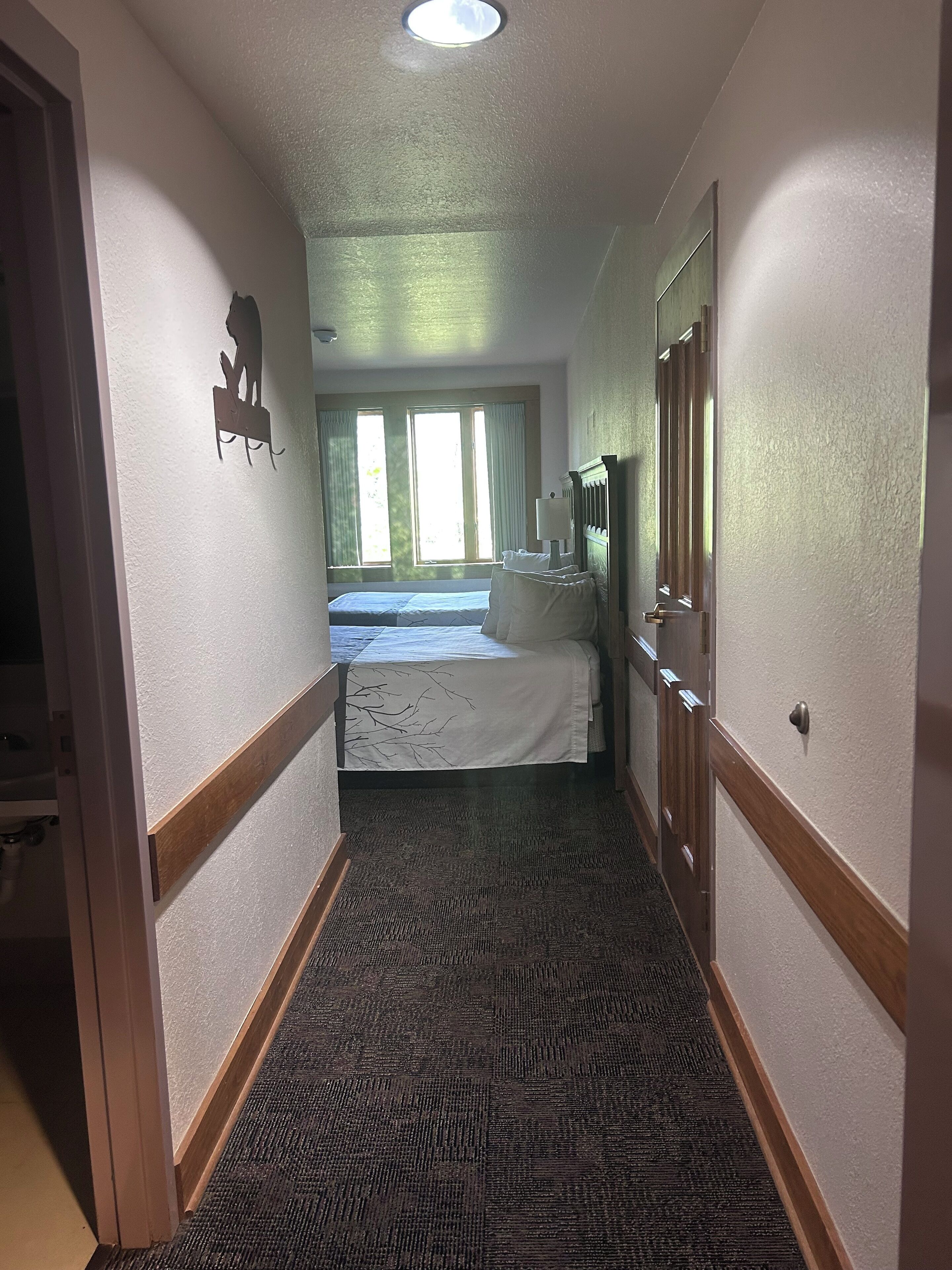 A hotel hallway leading to a room with a bed, a window, and a door.