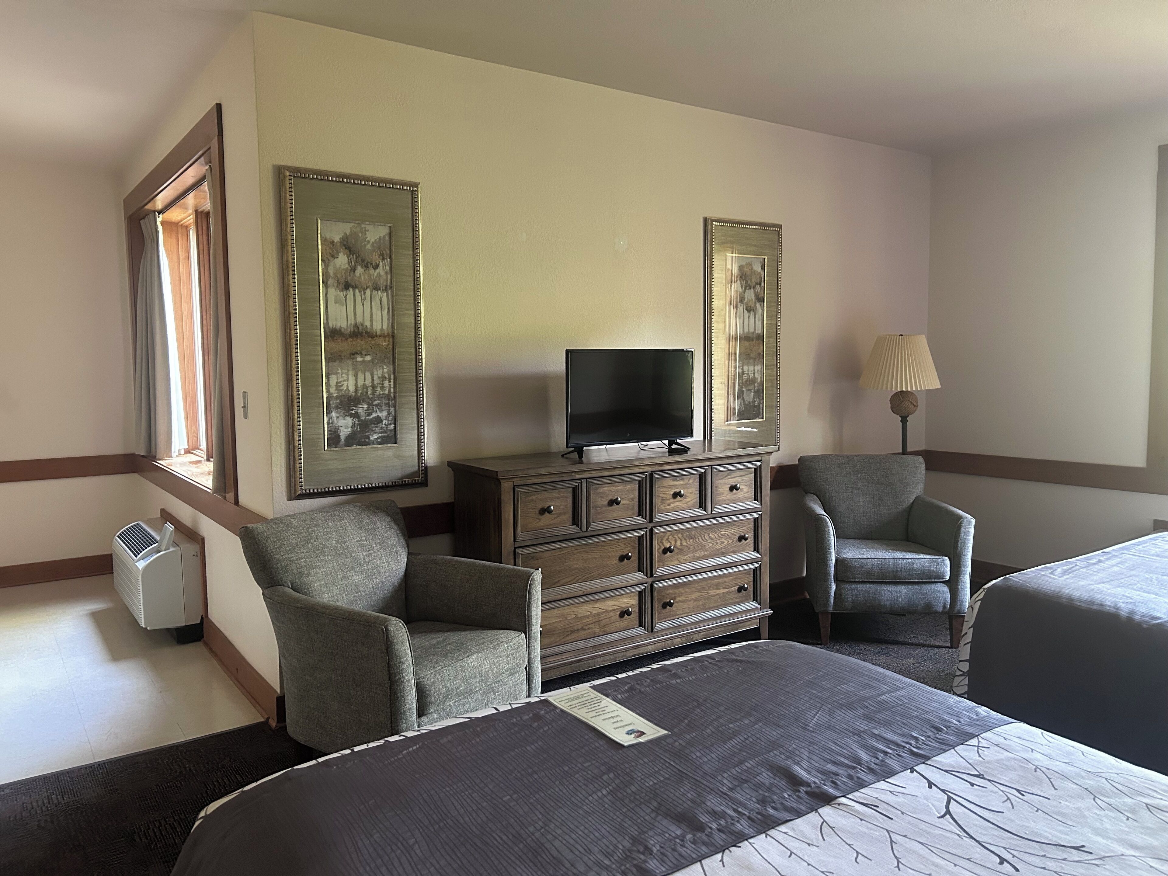 A hotel room with a bed, two armchairs, a dresser, a television, and two framed pictures on the wall.