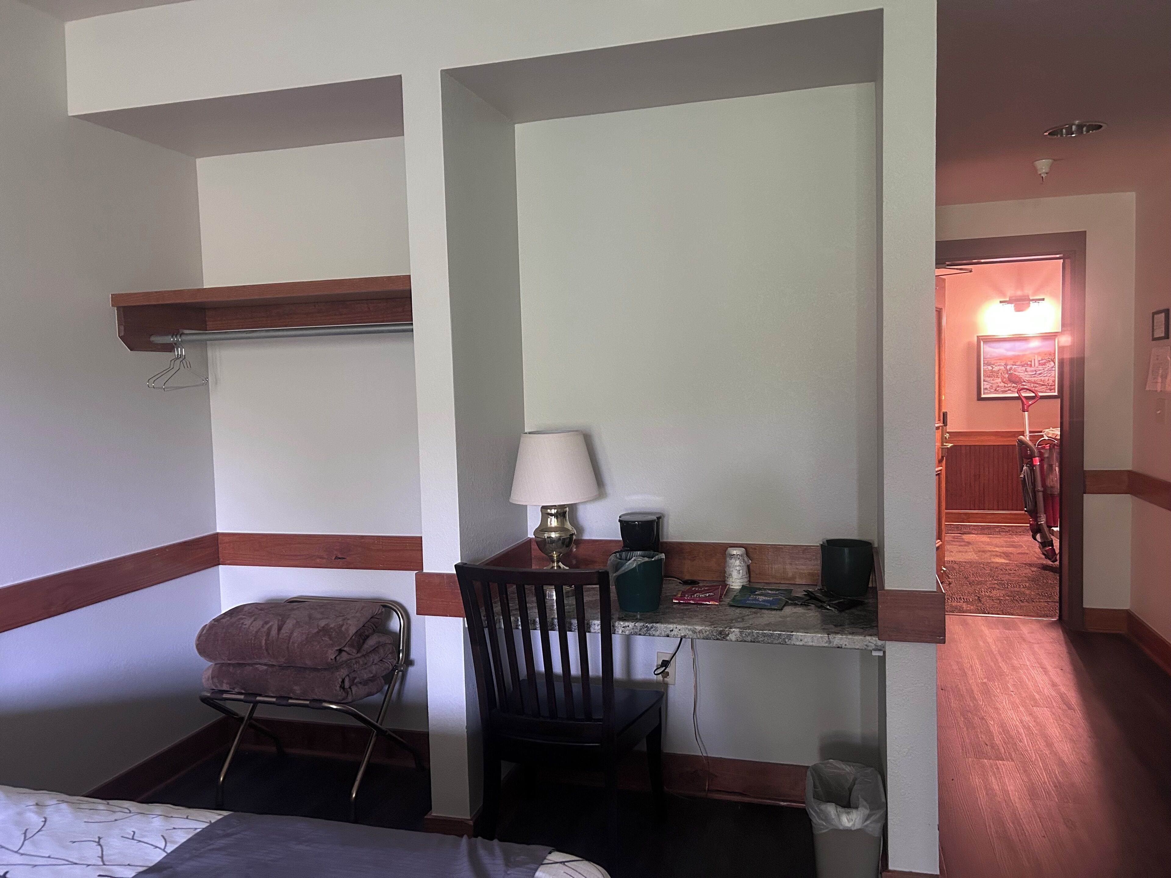 A hotel room with a bed, a desk with a lamp, a chair, a small table, a trash bin, and a vacuum cleaner in the hallway.