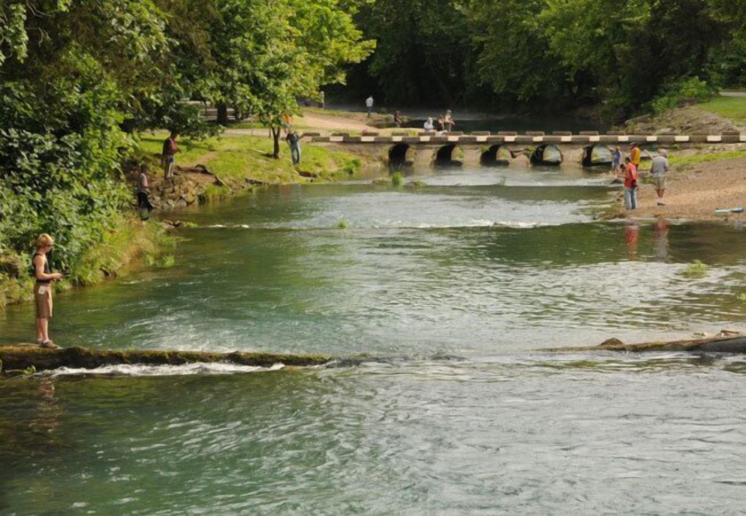 A river with people standing on the banks and a bridge in the background.