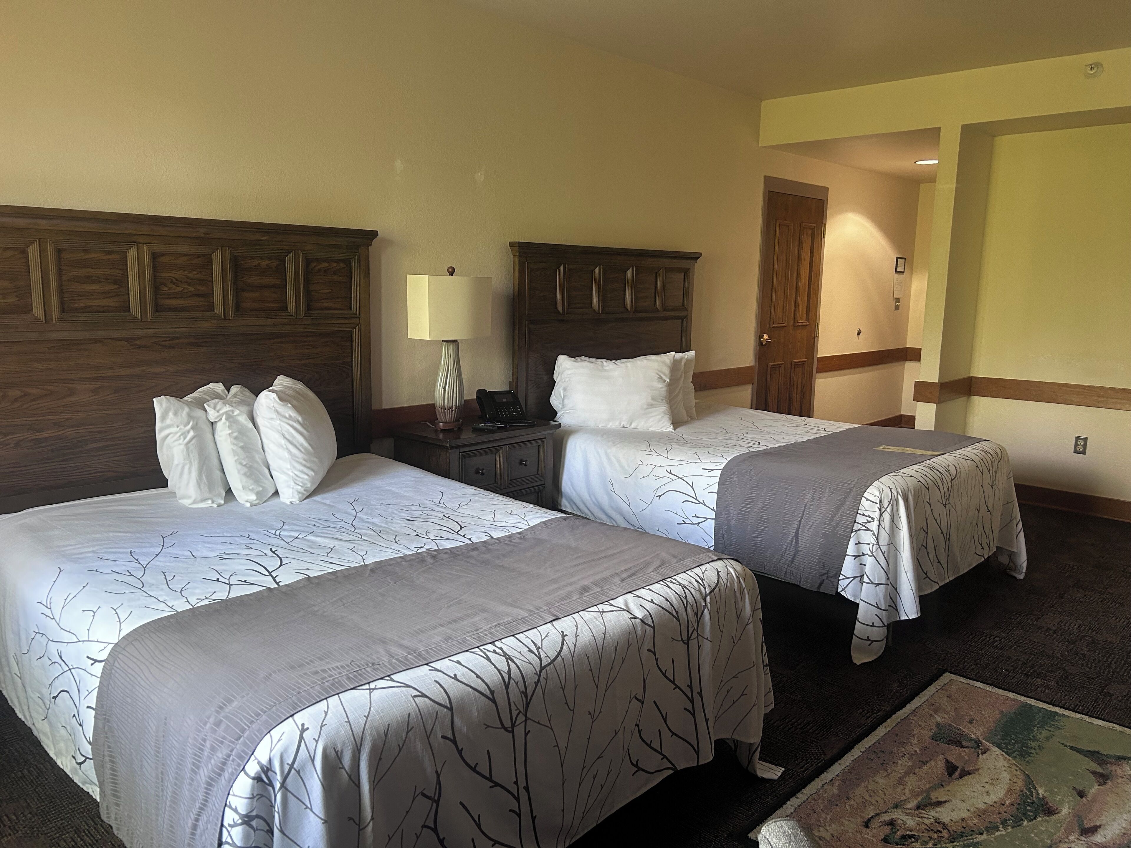 Une chambre d’hôtel avec deux lits, des têtes de lit en bois, une table de chevet avec une lampe et une porte donnant sur une autre pièce.