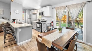 Una cocina moderna con una isla central, una zona de comedor con mesa de madera y vistas a un jardín trasero a través de amplios ventanales.