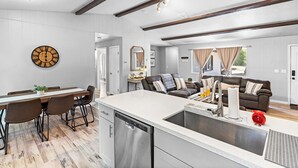 Una cocina moderna con lavavajillas, un comedor con mesa y sillas de madera, una sala de estar con sofá y mesa de centro, y un reloj de pared grande.