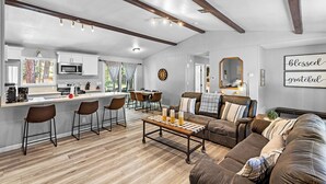 Una cocina moderna con armarios blancos, una isla central con taburetes, un comedor con mesa y sillas de madera, una sala de estar con sofá de cuero marrón y una mesa de centro, y una pared con un letrero que dice 'blessed grateful'.