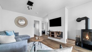 En moderne stue med sofa, sofabord og pejs. Der er et fjernsyn ophængt på væggen, og et træskab med en skål ovenpå.