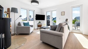 A spacious living room with a grey sofa, a coffee table, and a bookshelf. There is a flat-screen TV mounted on the wall and a large window providing a view of the outside.
