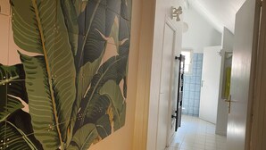 Ein Flur mit großem Wandbild eines tropischen Blattes, weiß gefliestem Boden und einer Tür zu einem weiteren Raum.