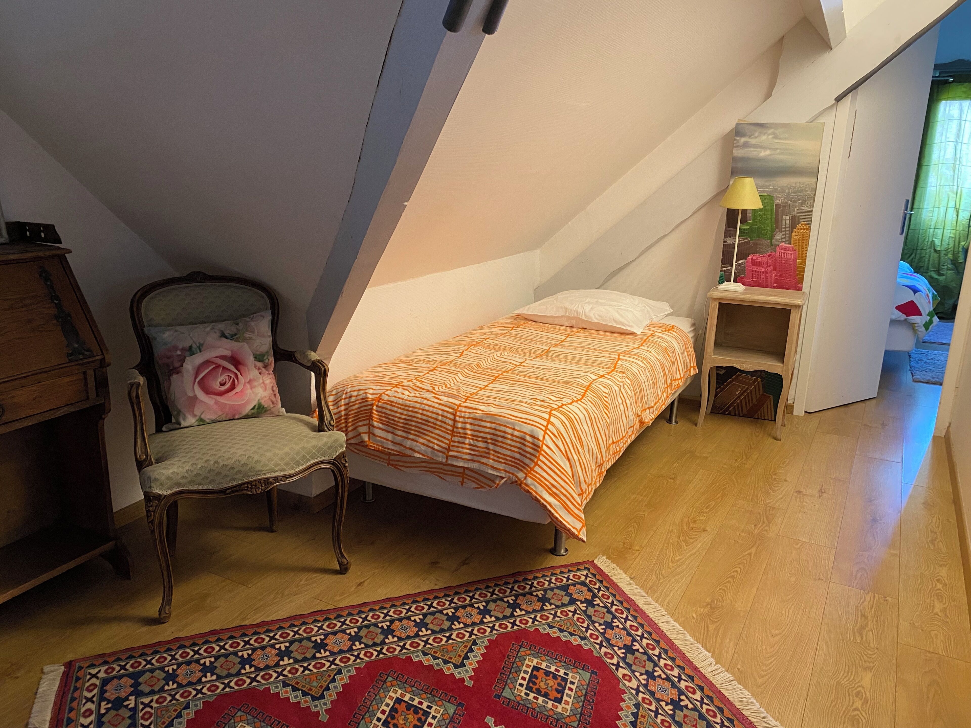 A small, cozy attic bedroom with a sloped ceiling, a single bed with an orange and white striped bedspread, a wooden chair with a floral cushion, a small bedside table with a lamp, and a patterned rug on the wooden floor.