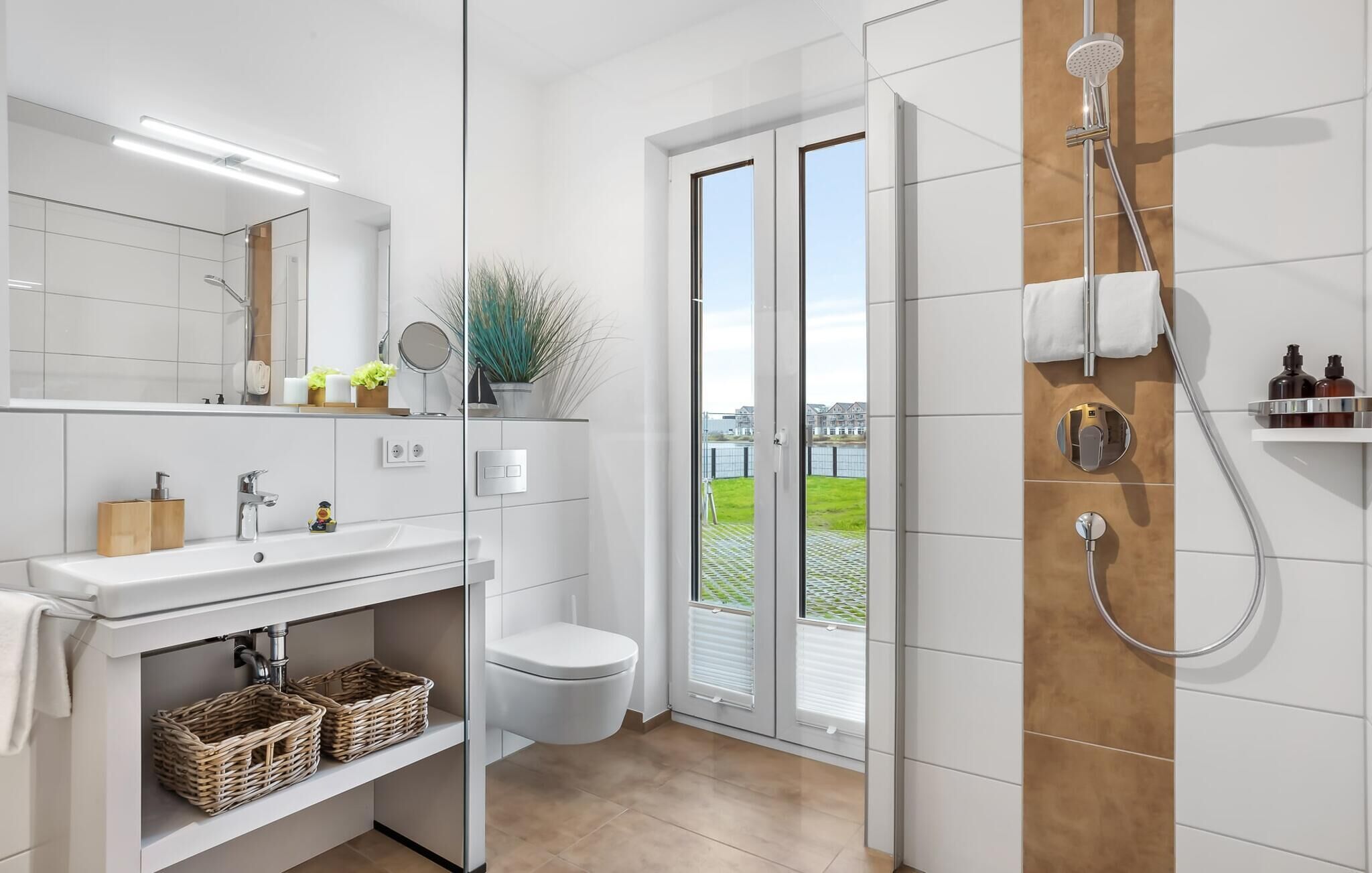 Un baño moderno con un espejo grande, baldosas blancas, un lavamanos, un inodoro y una ducha.