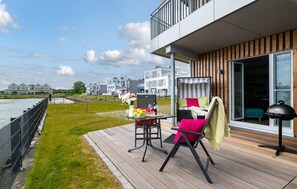 Une terrasse extérieure moderne, dotée d’une table et de chaises, offrant une vue sur un plan d’eau et des immeubles résidentiels.