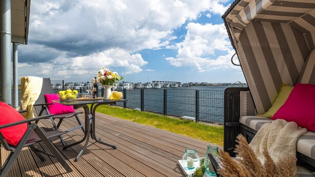A deck with a table set for two, a chair, and a sofa with cushions. The deck overlooks a body of water with buildings in the background. - Cozy apartment in Olpenitz with sauna (Olpenitz)
