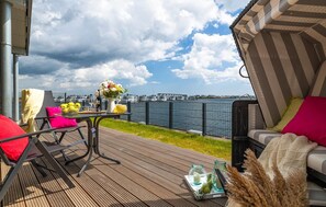 A deck with a table set for two, a chair, and a sofa with cushions. The deck overlooks a body of water with buildings in the background. - Cozy apartment in Olpenitz with sauna (Olpenitz)