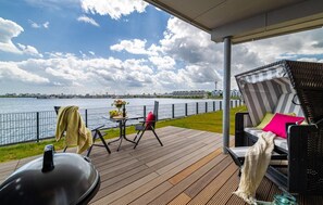 Une terrasse équipée d’un barbecue, de chaises et d’une table agrémentée de fleurs, offrant une vue sur une étendue d’eau et des bâtiments.