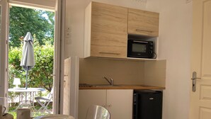 Eine kompakte Küche mit Mikrowelle, Spüle und Kühlschrank. Vor dem Fenster steht ein Esstisch mit Stuhl und ein kleiner Sonnenschirm.