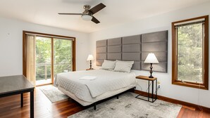 Un dormitorio con una cama grande, un ventilador de techo, dos lámparas de noche, una mesa de madera y vistas al exterior a través de una puerta corrediza de vidrio.