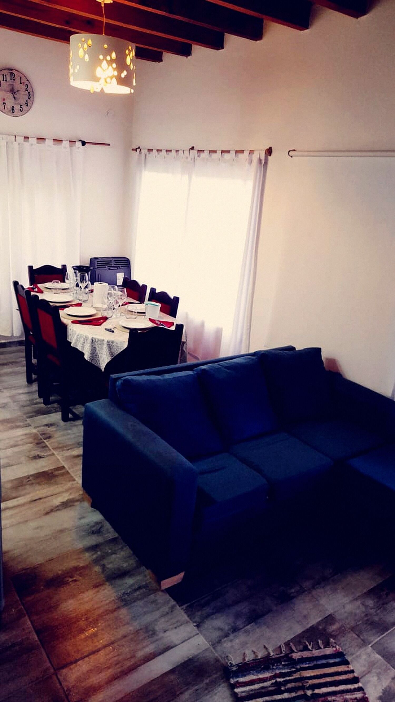 Un comedor con una mesa preparada para cuatro personas, un sofá y un reloj en la pared.