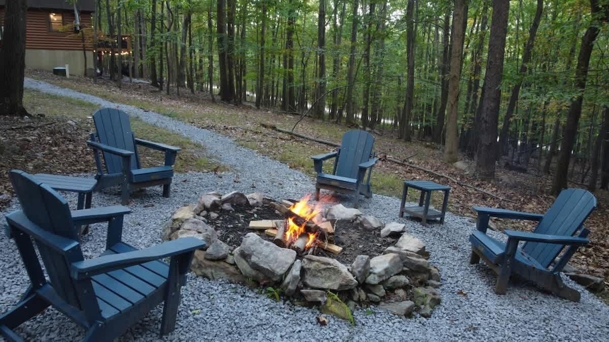 Ein Feuerkorb mit vier Adirondack-Stühlen, die darum platziert sind, inmitten eines bewaldeten Geländes mit einem Kiesweg, der zu einem Gebäude führt.