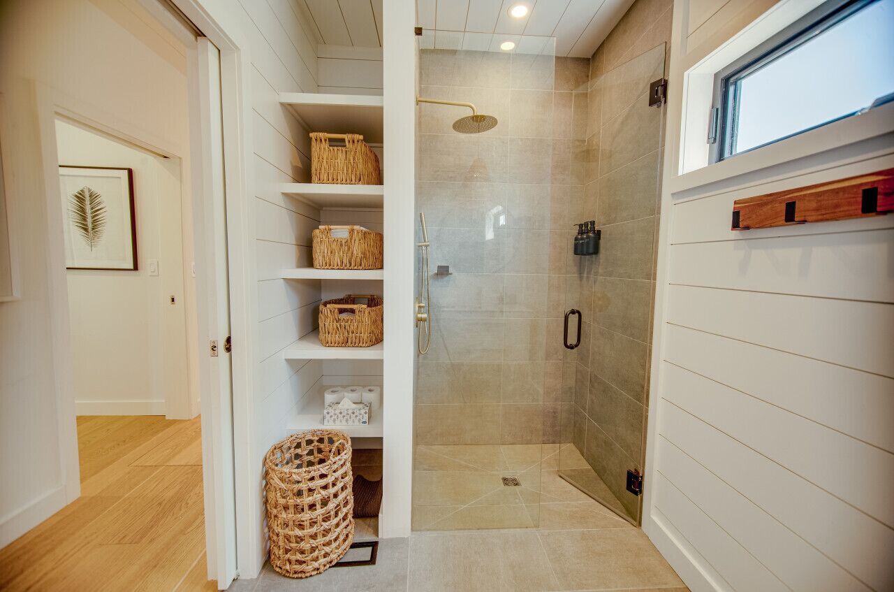 A walk-in shower with a glass door, a wooden shelf with baskets, and a window above the shower.