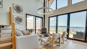 A spacious living room with a large window overlooking a beach, a fireplace, and a wooden staircase. - Zen Oceanfront Chalet- Hot Tub & Sauna -  Private (South Shore)