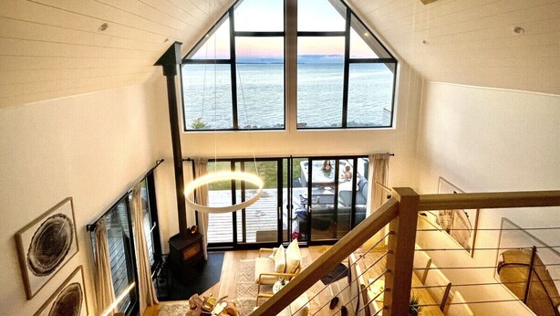 A high-ceilinged room with large windows overlooking a body of water, a staircase, and a hanging light fixture. - Zen Oceanfront Chalet- Hot Tub & Sauna -  Private (South Shore)