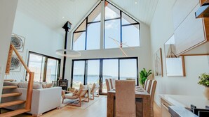 A spacious living area with a high ceiling, large windows, a dining table with chairs, and a staircase leading to another level. - Zen Oceanfront Chalet- Hot Tub & Sauna -  Private (South Shore)