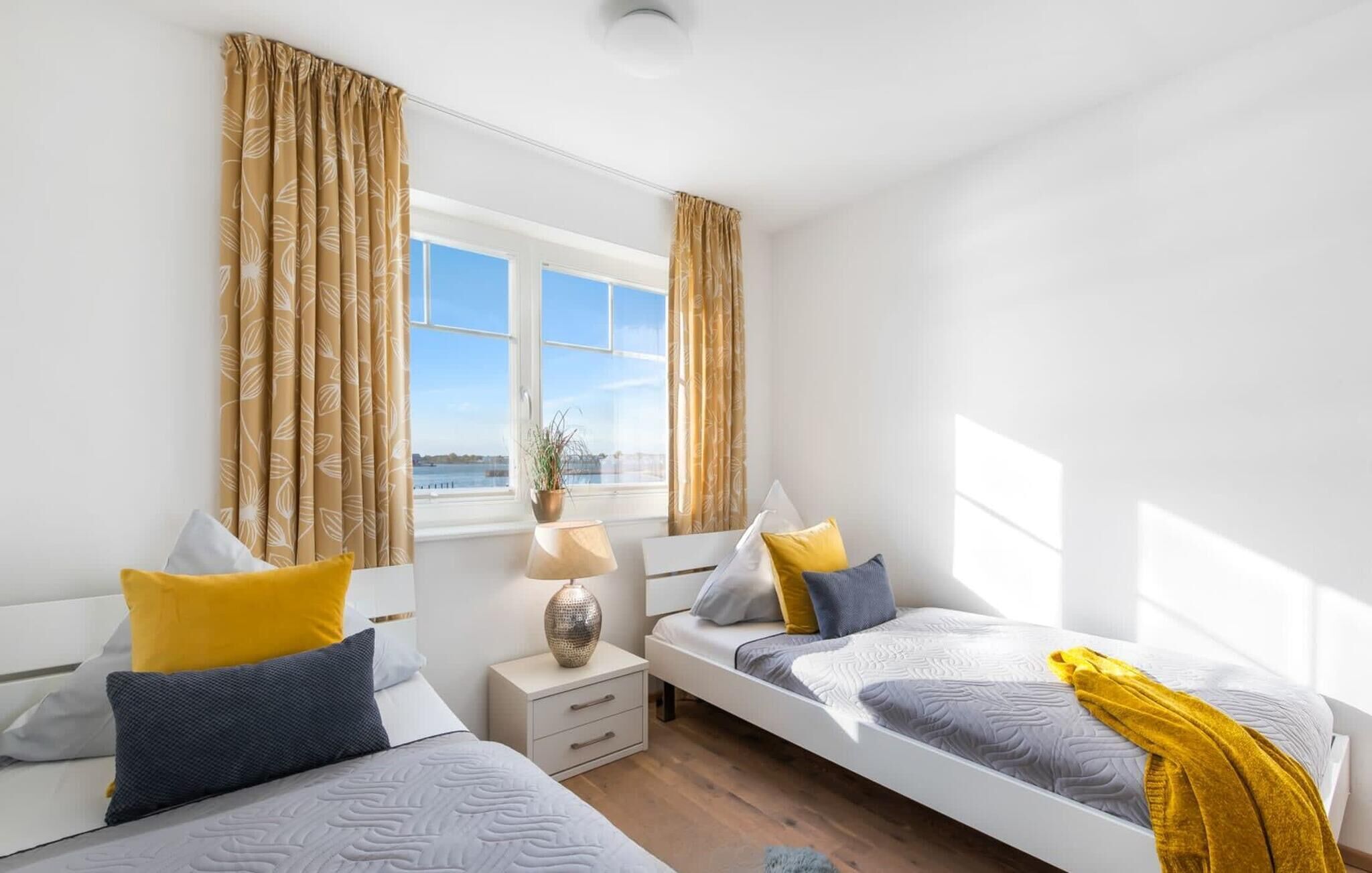 A modern bedroom with two beds, a bedside table, a lamp, and a window with curtains.