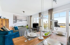 Un comedor moderno con una mesa de madera dispuesta para una comida, un televisor al fondo y vistas al mar a través de amplias ventanas.