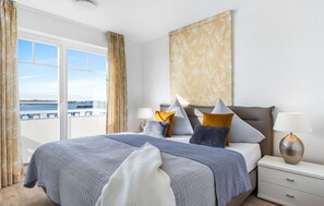 Habitación de hotel con una cama grande, dos mesitas de noche, vista al mar y una ventana con cortinas.