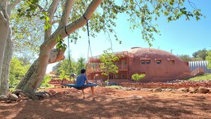 A person sitting on a swing under a tree, a unique geodesic dome building in the background, and a clear blue sky.