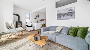 Un salon moderne avec un canapé gris, une table basse en bois et un fauteuil noir. On y trouve une cheminée, une plante suspendue et un tableau encadré au mur.