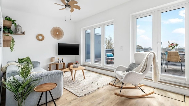 Un salon moderne avec un canapé gris, un fauteuil à bascule blanc, un téléviseur à écran plat et de grandes fenêtres donnant sur l’extérieur.