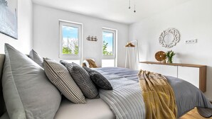 Une chambre moderne avec un lit, deux fenêtres, un ventilateur de plafond, un miroir et une table de chevet sur laquelle se trouve un vase.