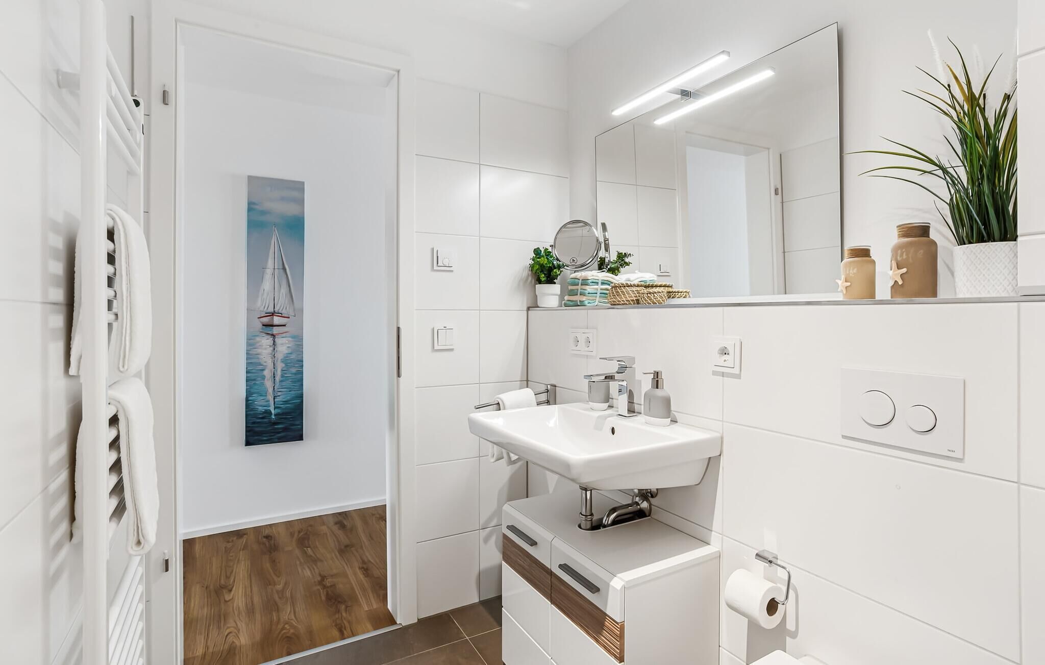 A modern bathroom with a white sink, mirror, and towel rack. There is a painting of a sailboat on the wall, and a plant on the counter.