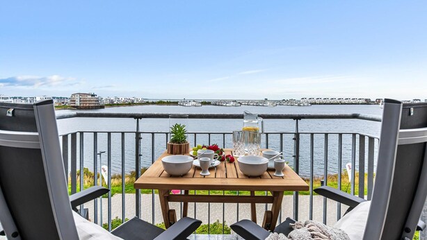 A dining table with bowls, cups, and a pitcher, set on a balcony with a view of a waterfront and buildings in the background. - Nice apartment in Olpenitz with WiFi (Olpenitz)