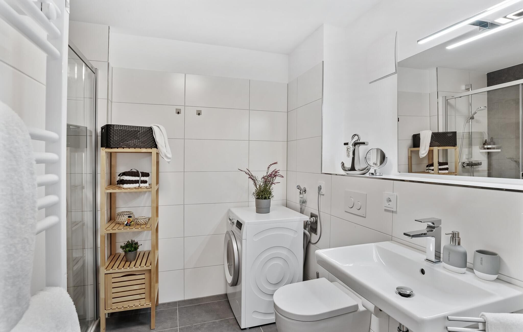 Un baño moderno con baldosas blancas, lavadora, inodoro, lavamanos con espejo y estante con toallas y planta.