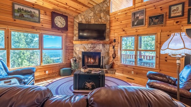 Interior acogedor de una cabaña con chimenea de piedra, sofás de cuero y ventanas amplias que ofrecen vistas al exterior.