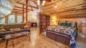 Interior de una cabaña de troncos con techo de madera, una cama con colcha, un sofá, una mesa de centro y vistas al exterior.
