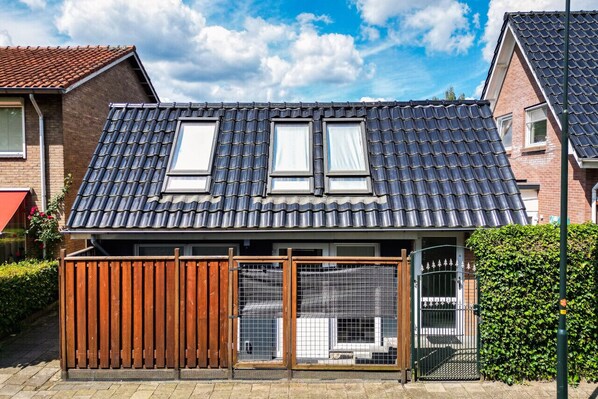 A residential house with a dark tiled roof, two windows, a wooden fence, and a metal gate.