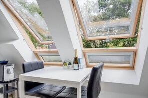 Una habitación moderna con una mesa blanca, sillas de cuero negro y una botella de champán. Hay tragaluces y se ve arboles desde el interior.