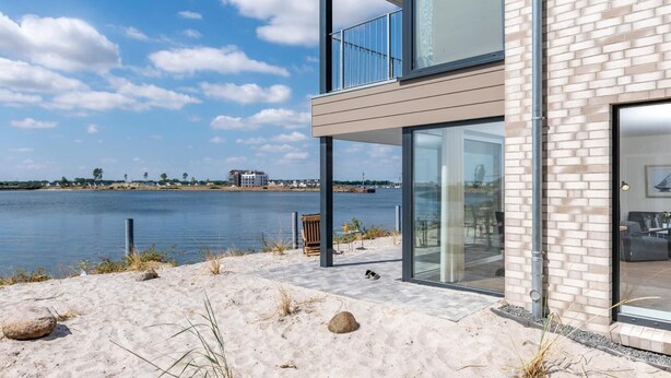 A modern building with a glass balcony overlooking a body of water, a sandy area with rocks, and a distant view of a town. - Lovely apartment in Olpenitz with sauna (Olpenitz)