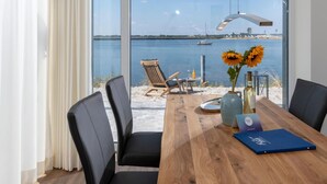 A modern room with a large window overlooking a beach and sea. There is a wooden table with chairs, a vase with sunflowers, and a bottle. - Lovely apartment in Olpenitz with sauna (Olpenitz)
