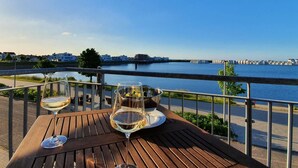 Eine Holzterrasse mit zwei Weingläsern und einer Metallkanne, mit Blick auf einen ruhigen Fluss mit Gebäuden und einer Brücke in der Ferne.