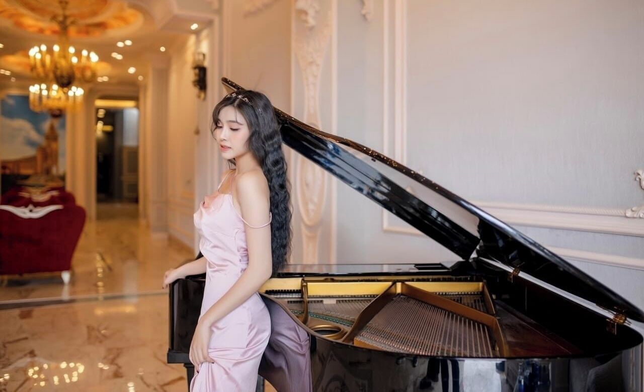 A woman in a pink dress sitting at a grand piano in a luxurious hallway with ornate decorations and a chandelier.