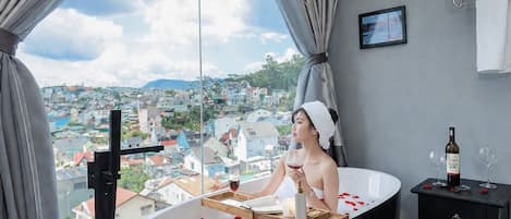 Una habitación con bañera, ventana con cortinas, un perchero y una pequeña mesa con una botella y vasos.