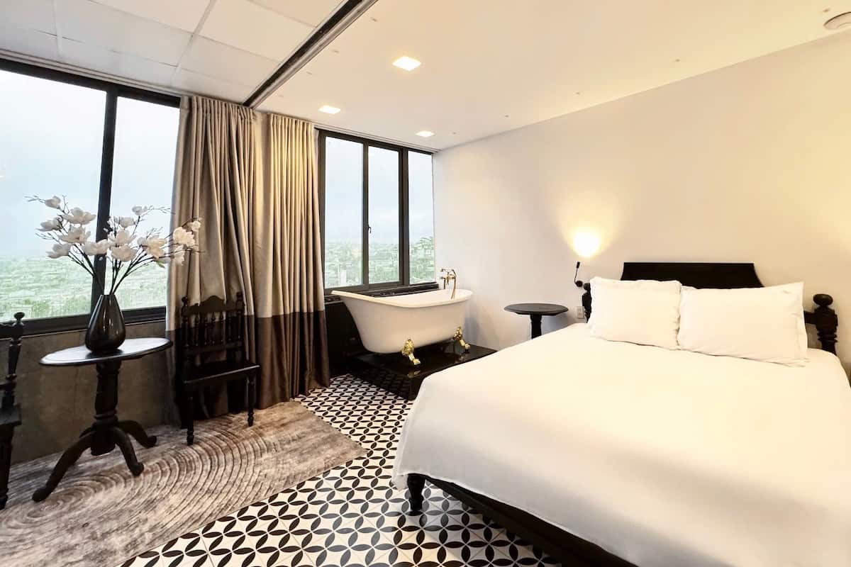 A hotel room with a large bed, a bathtub, a round table, and a view of the city through large windows.