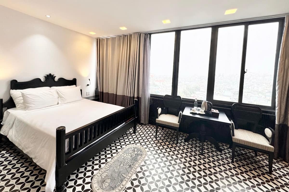 A hotel room with a bed, a desk with a chair, a view of the city through large windows, and a patterned floor.