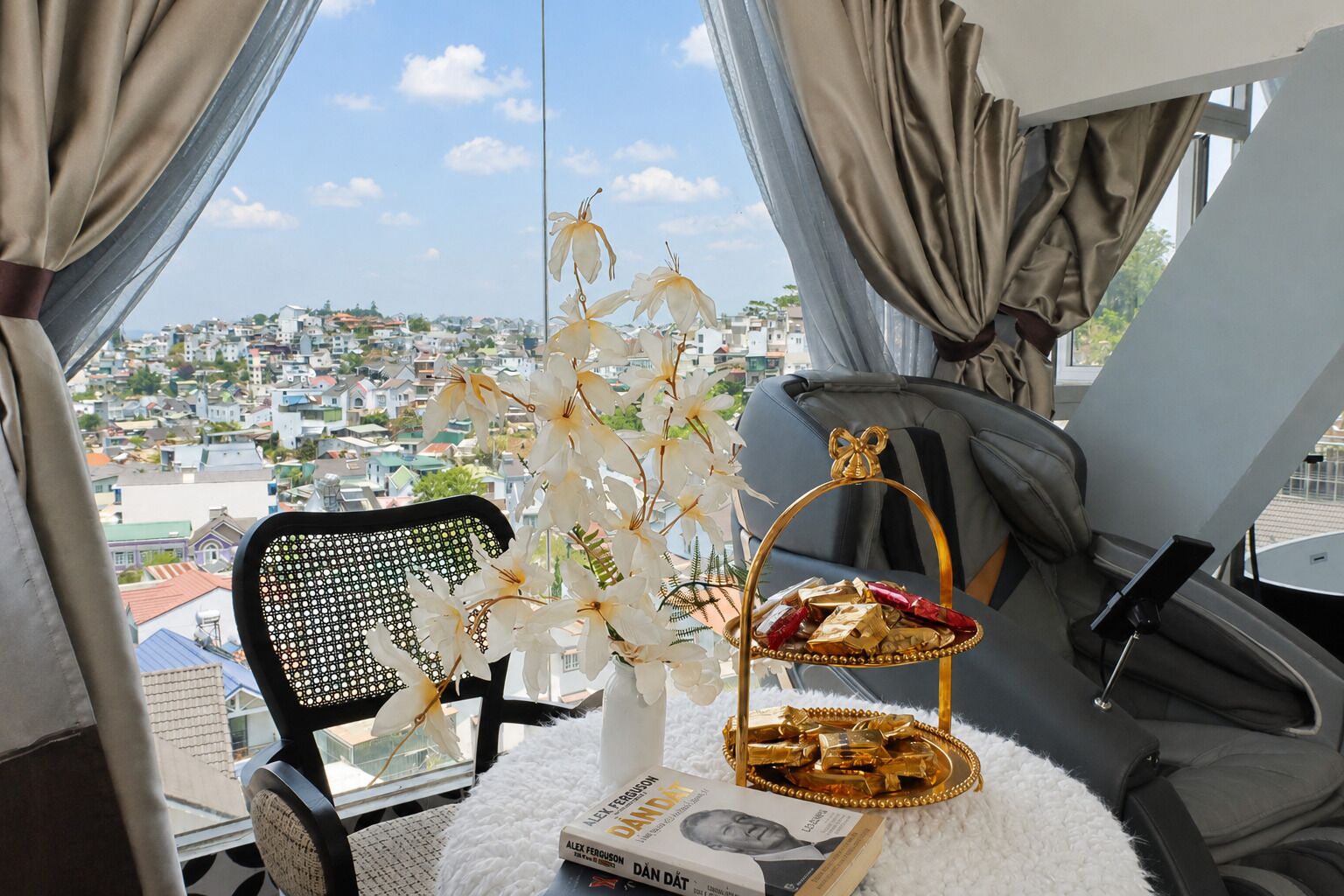 A room with a view of a cityscape, a black chair, a white vase with flowers, a tray with chocolates, and a book titled 'DAN DÂT'.