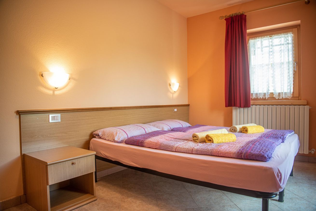 Un lit simple avec une tête de lit, une table de chevet, une lampe fixée au mur, une fenêtre avec un rideau et un radiateur.
