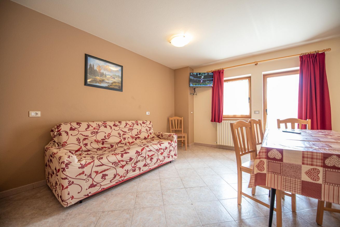 Ein Wohnzimmer mit einer Couch, einem Esstisch mit Stühlen, einem an der Wand montierten Fernseher und einem Fenster mit Vorhängen.