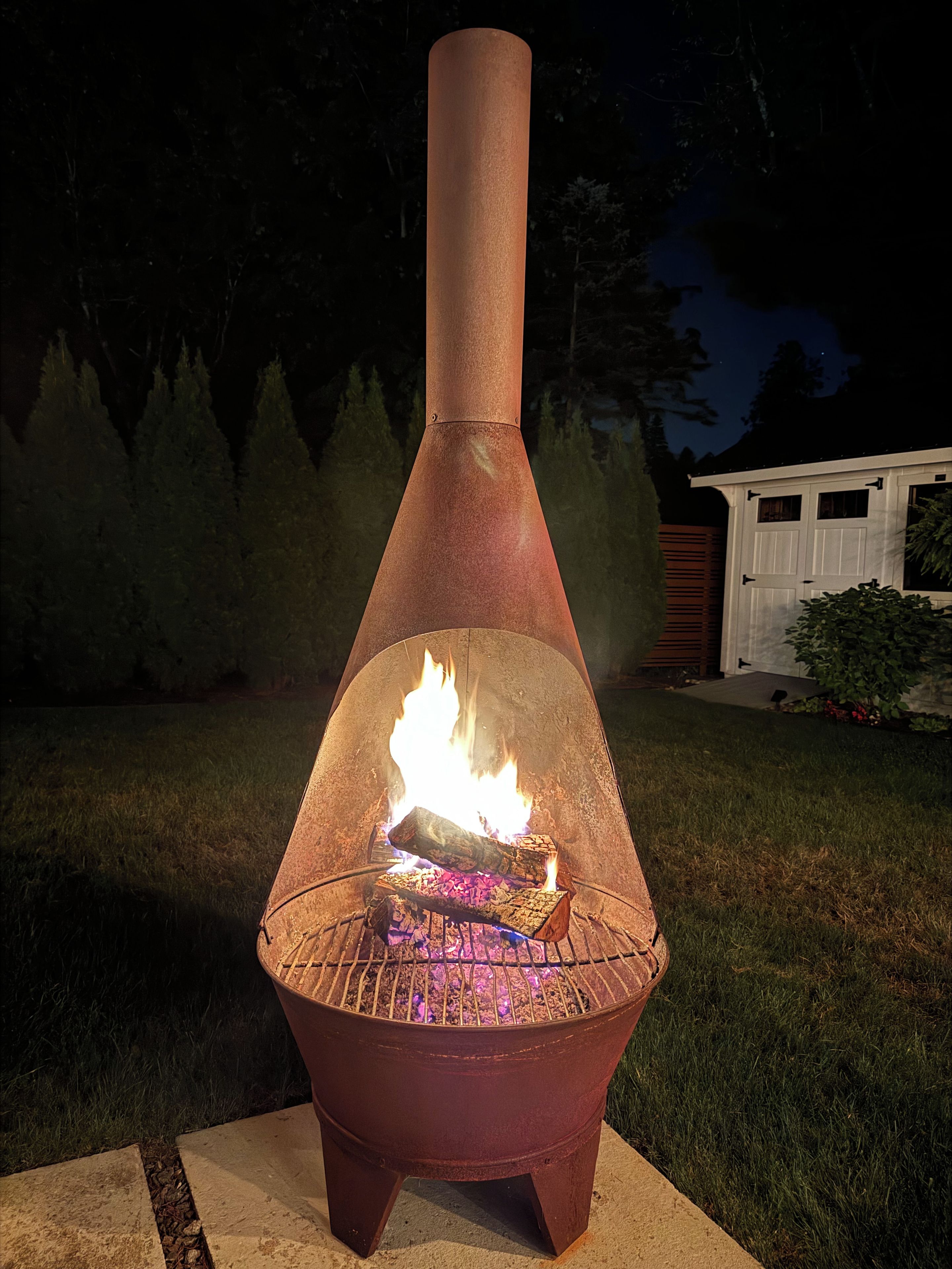 A metal fire pit with a chimney, burning wood, and a white shed in the background.